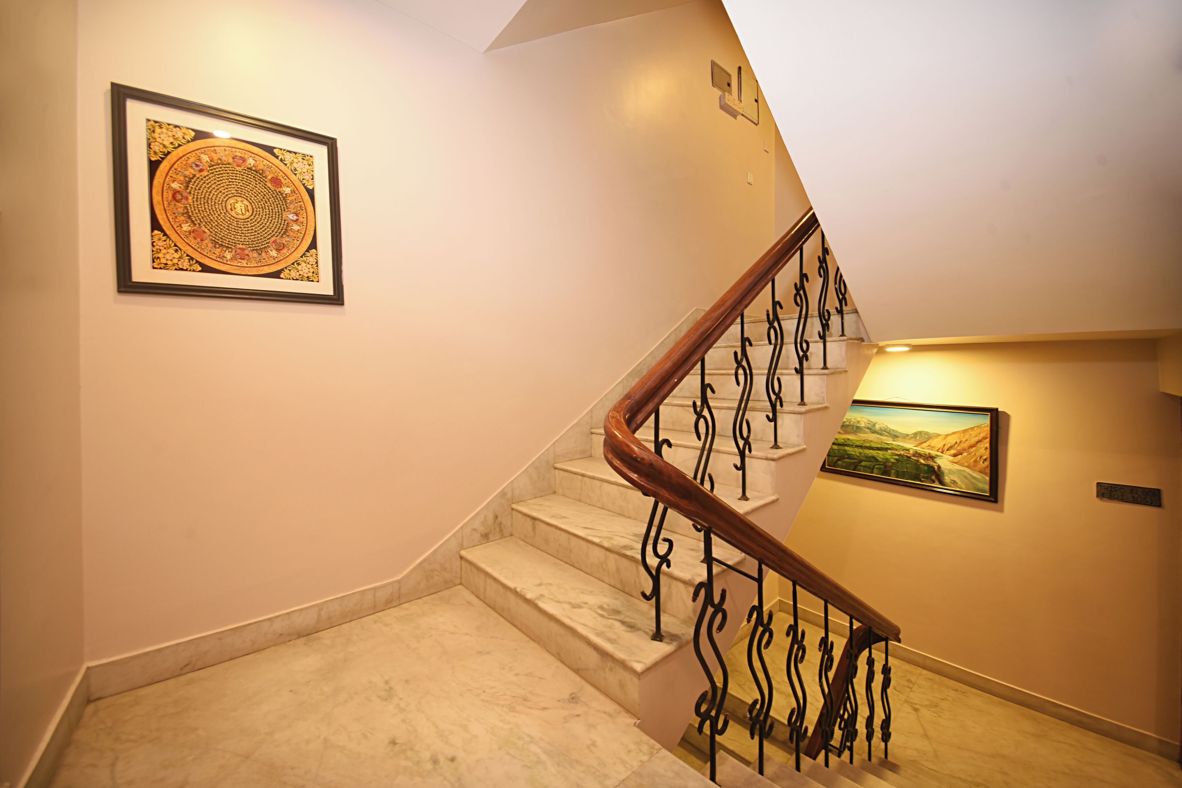 A marble staircase with a wooden handrail and black wrought iron balusters, leading to a landing with a framed artwork and a landscape painting.