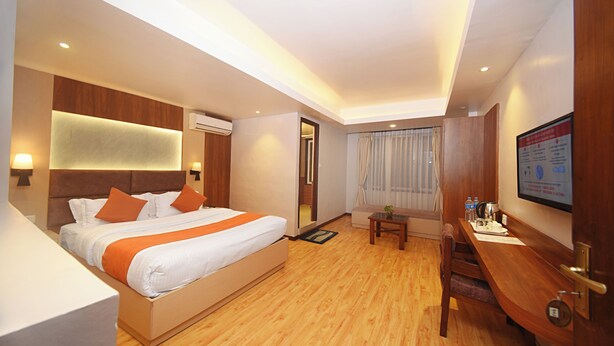 A hotel room with a large bed, wooden flooring, a desk with a chair, a small table with a plant, and a wall-mounted TV. - Hotel Mums Home (Kathmandu)