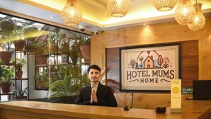 A hotel reception desk with a wooden counter, a framed logo above it, and potted plants in the background. - Hotel Mums Home (Kathmandu)