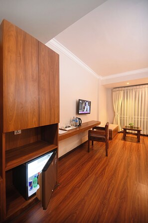 A compact hotel room with a wooden wardrobe, a small fridge, a television, a wooden desk, a chair, and a window with curtains. - Hotel Mums Home (Kathmandu)