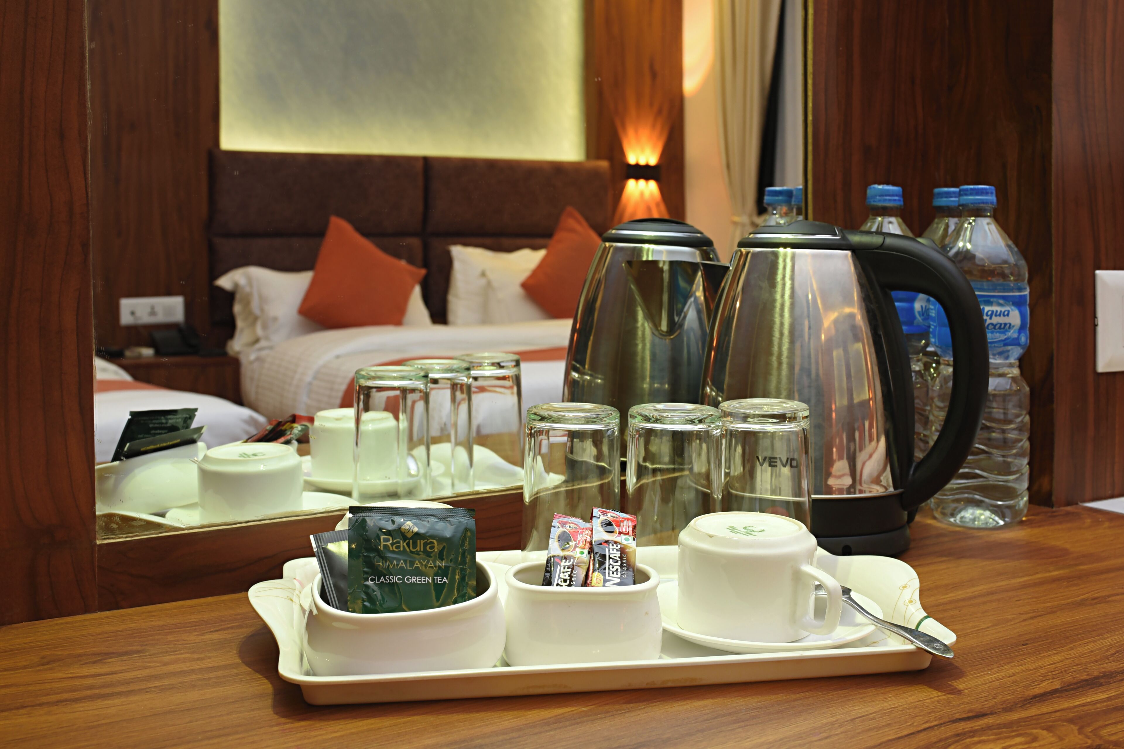 A hotel room with a wooden headboard, a bed with orange pillows, a bedside table with a phone, a tray with tea, coffee, and snacks, and a water bottle.