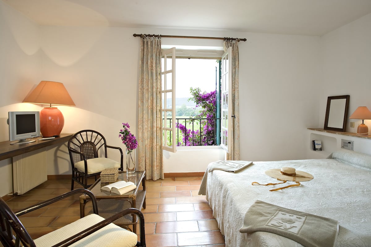 Un dormitorio con una cama, una silla, un escritorio con lámpara, un televisor y una ventana con cortinas.