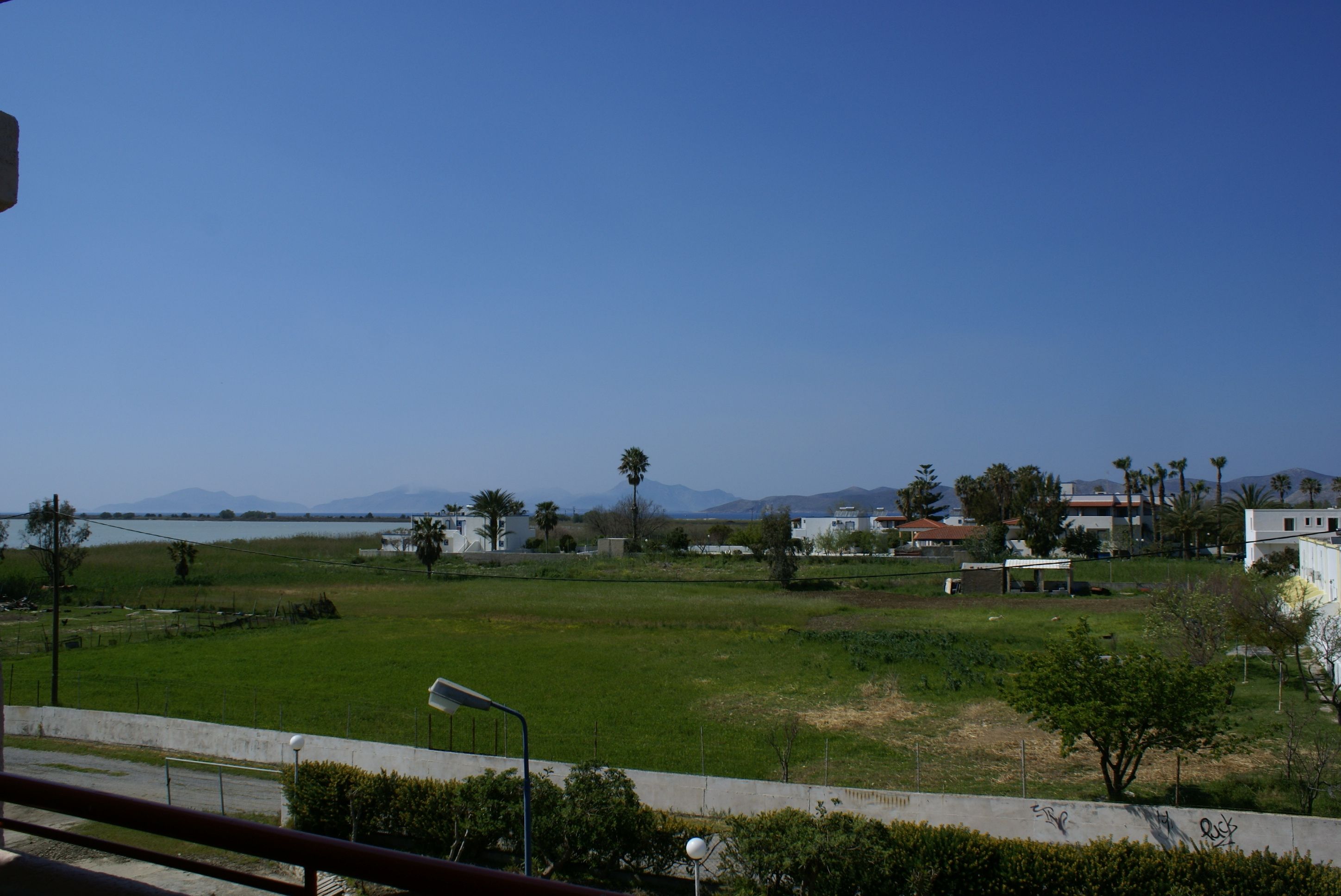 A view of a coastal area with a grassy field, a few buildings, and mountains in the distance.