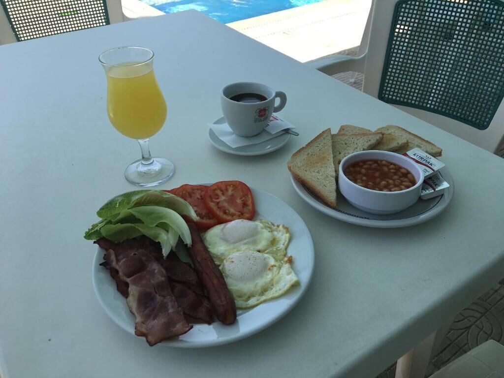 Ein Frühstückstisch mit Eiern, Speck, Tomaten, einem Glas Orangensaft, einer Tasse Kaffee, Toast und einer Schüssel Bohnen.