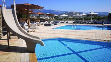 Una piscina con tobogán, sillas de descanso, sombrillas y montañas al fondo.