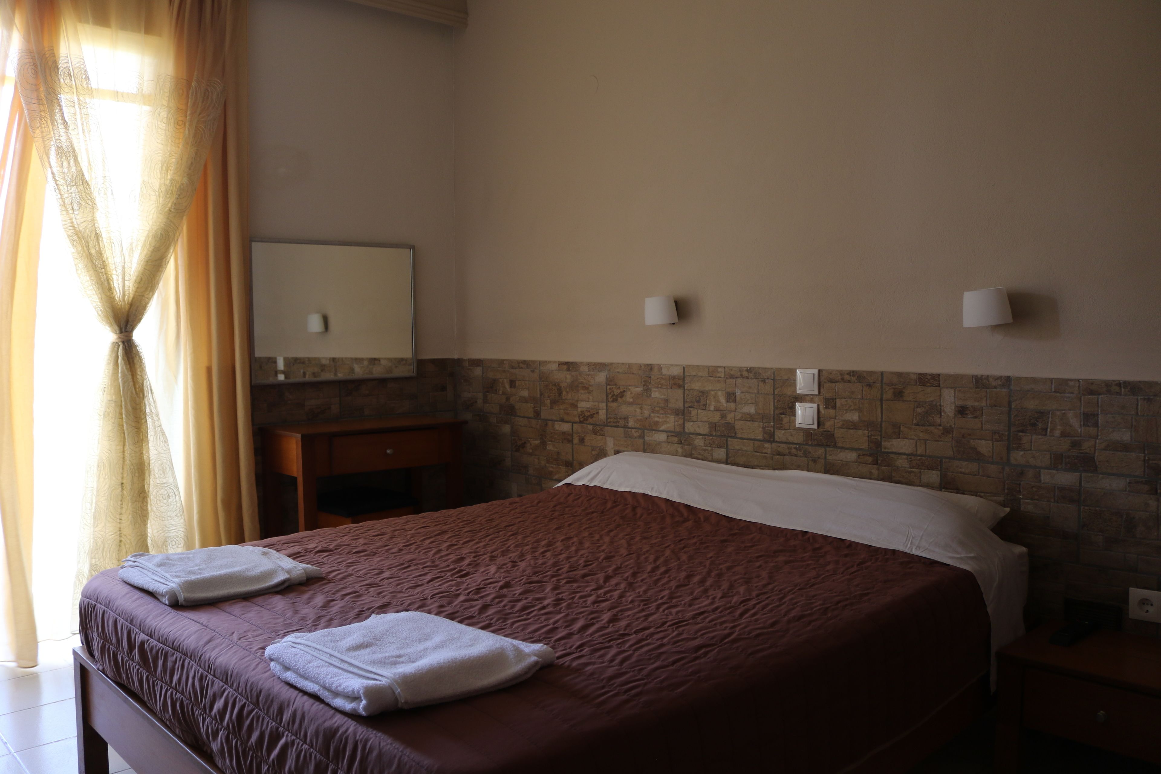 Quarto de hotel com uma cama grande, uma mesa de cabeceira de madeira, um espelho e uma parede com padrão de pedra.