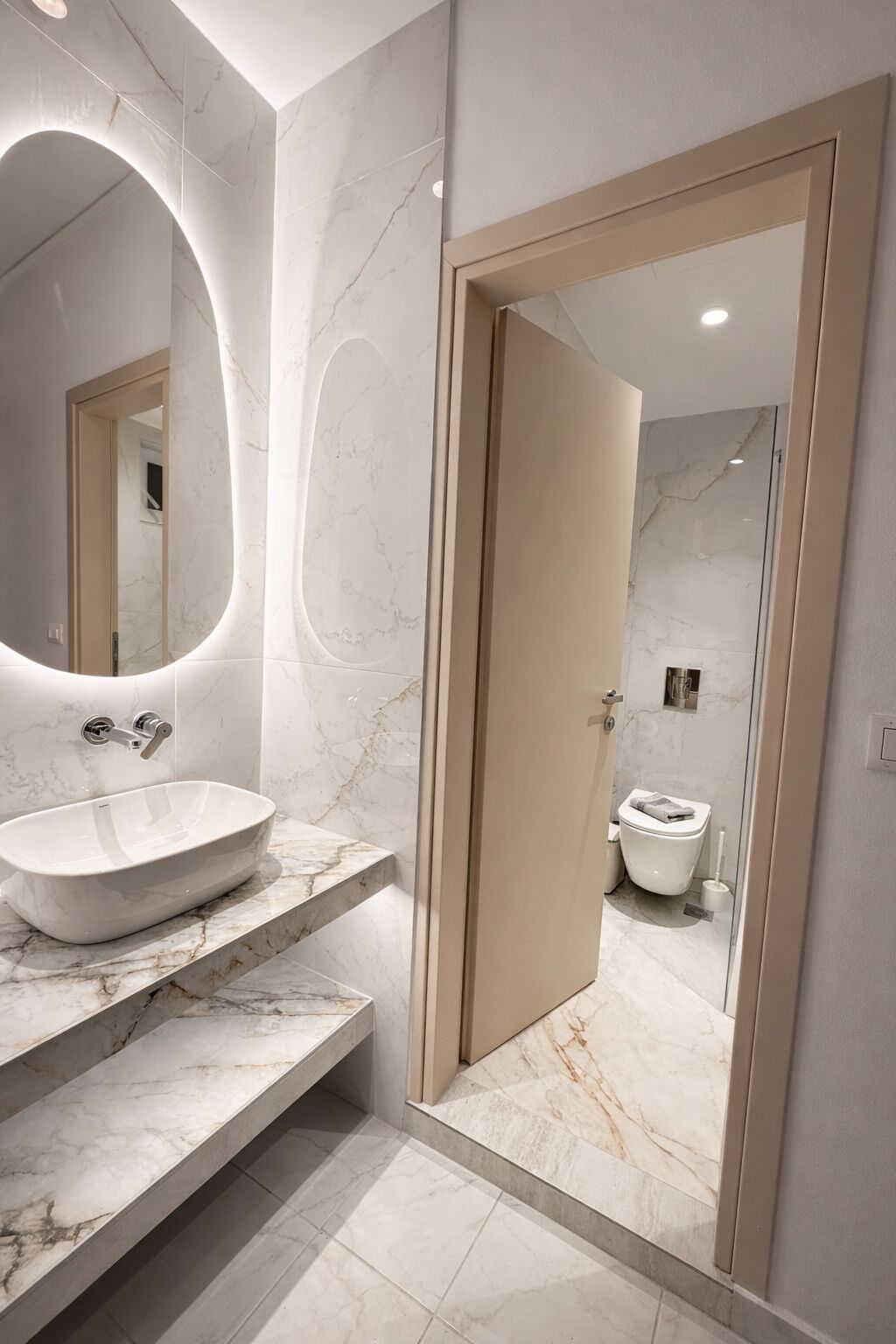Un baño moderno con lavamanos de mármol, un espejo grande y un inodoro visible a través de una puerta abierta.