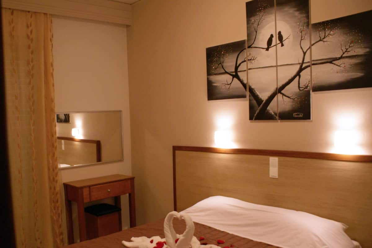 Habitación de hotel con cama, mesita de noche, espejo y un cuadro colgado en la pared que muestra pájaros en un árbol.