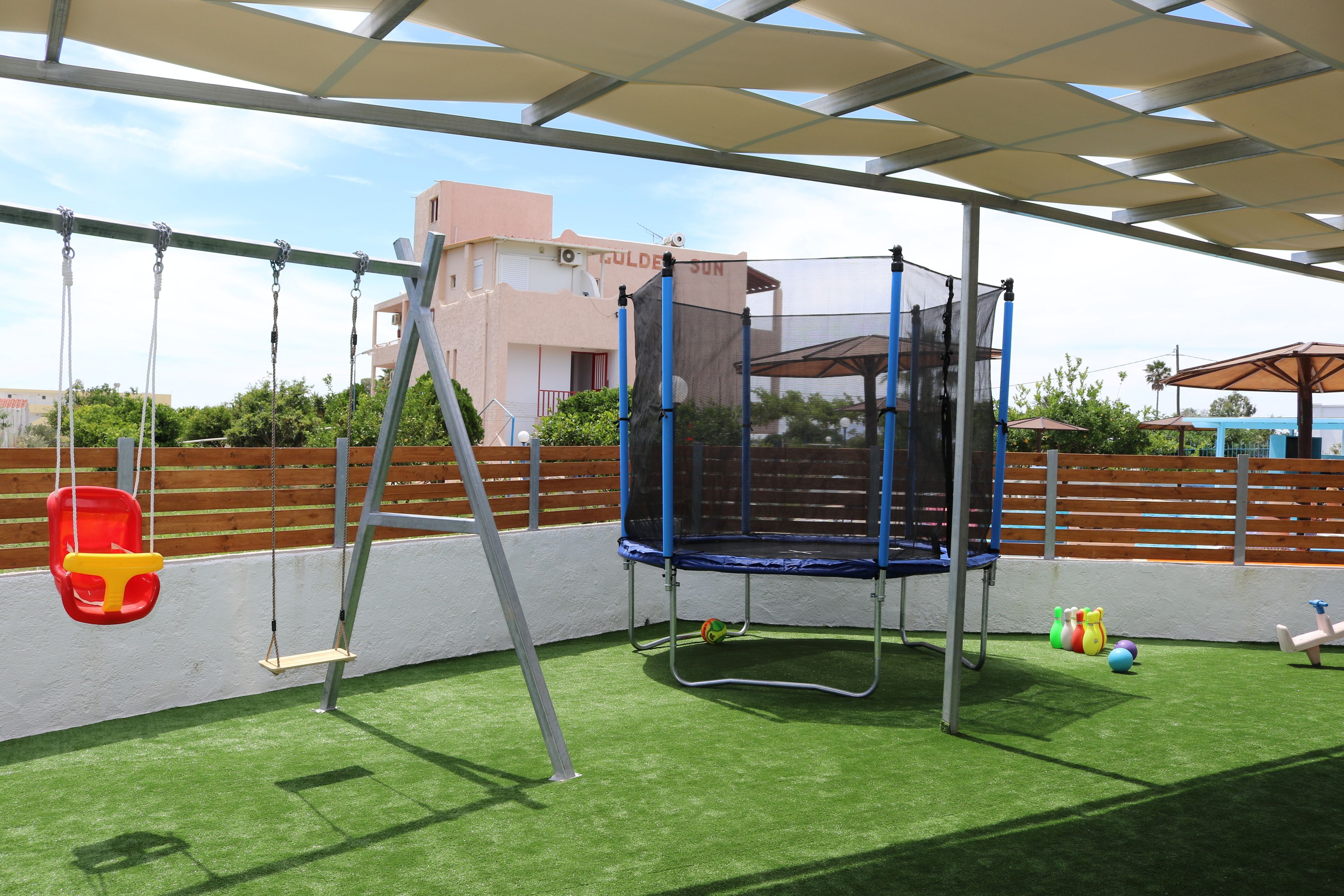 A playground with swings, a trampoline, and a climbing structure under a shaded area.