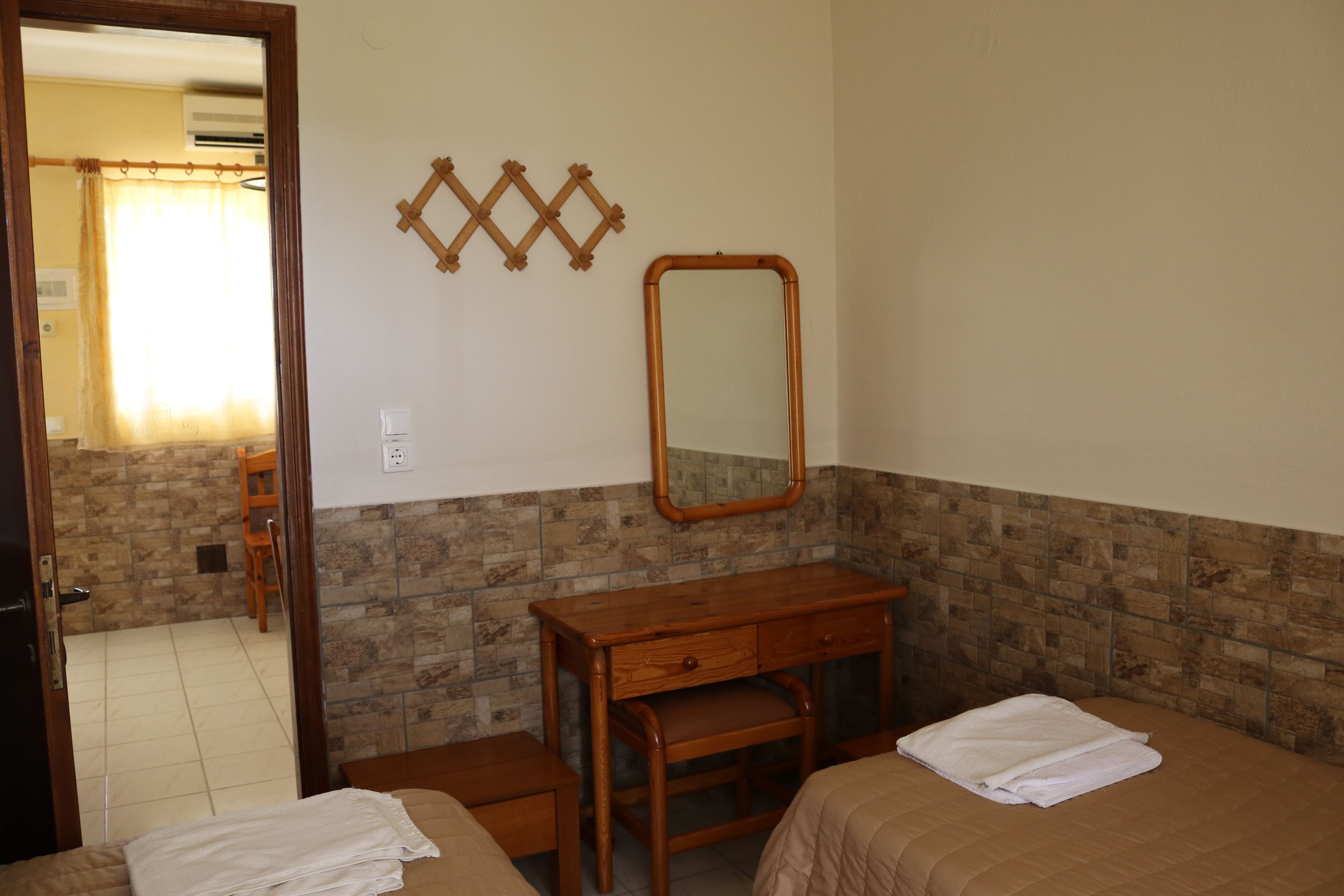 Una camera d'albergo con un comò in legno, un letto con coperta bianca, uno specchio a parete e un bagno visibile attraverso una porta aperta.