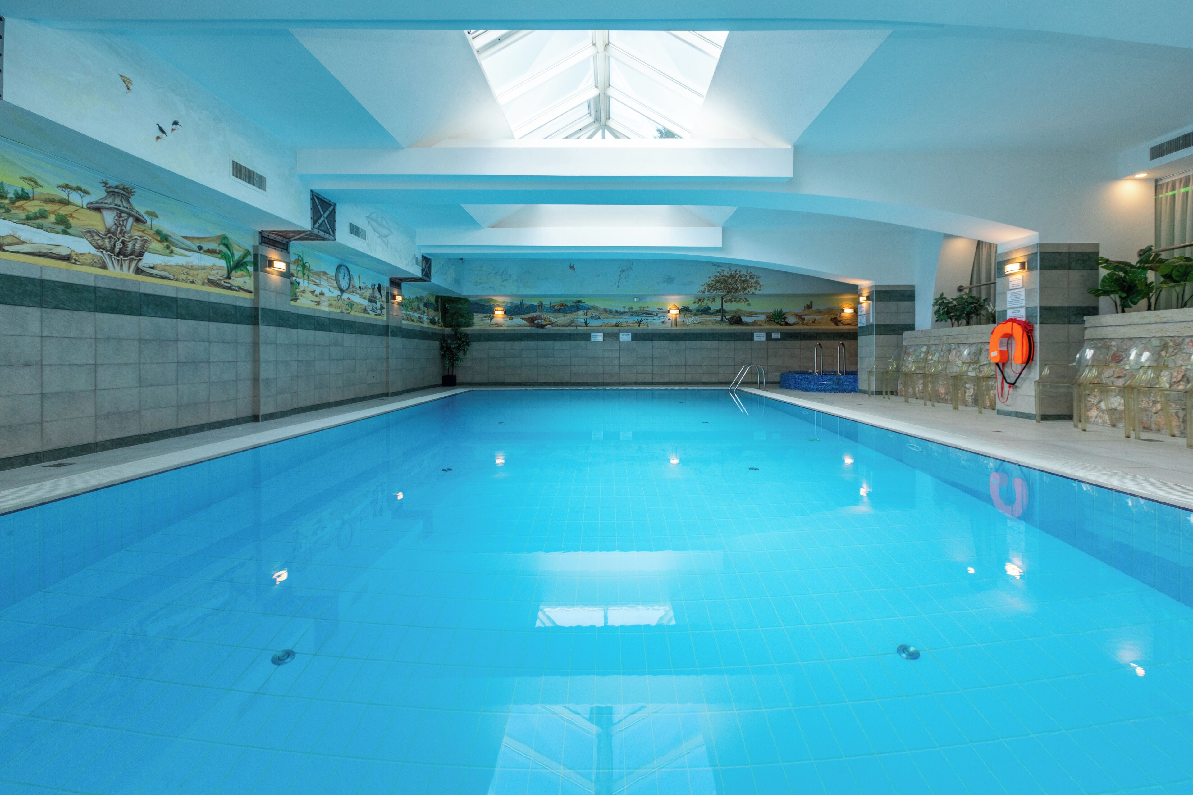 An indoor swimming pool with a mural on the walls, a lifebuoy, and a skylight.
