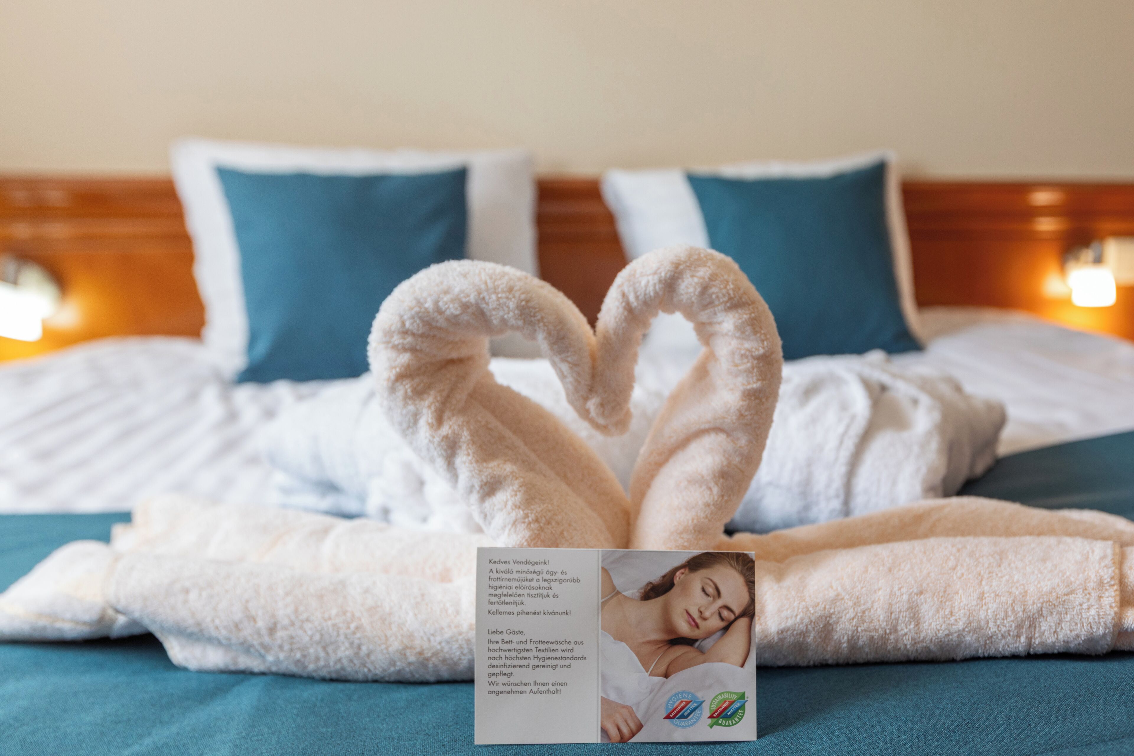 Una cama bien hecha con una toalla dispuesta en forma de corazón, un folleto y una colcha color aguamarina.