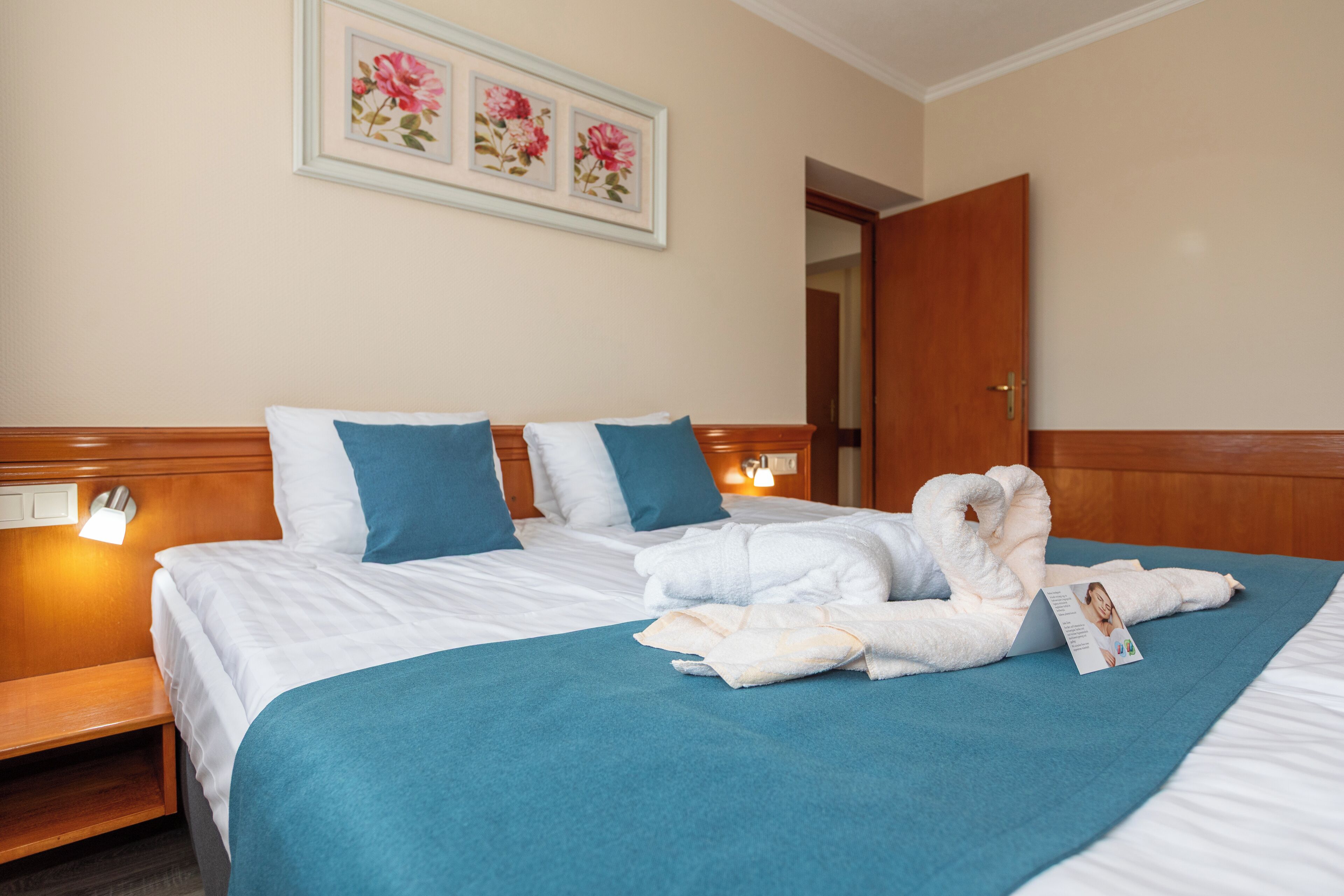 A hotel room with a bed, pillows, a blue blanket, a towel, and a framed picture on the wall.