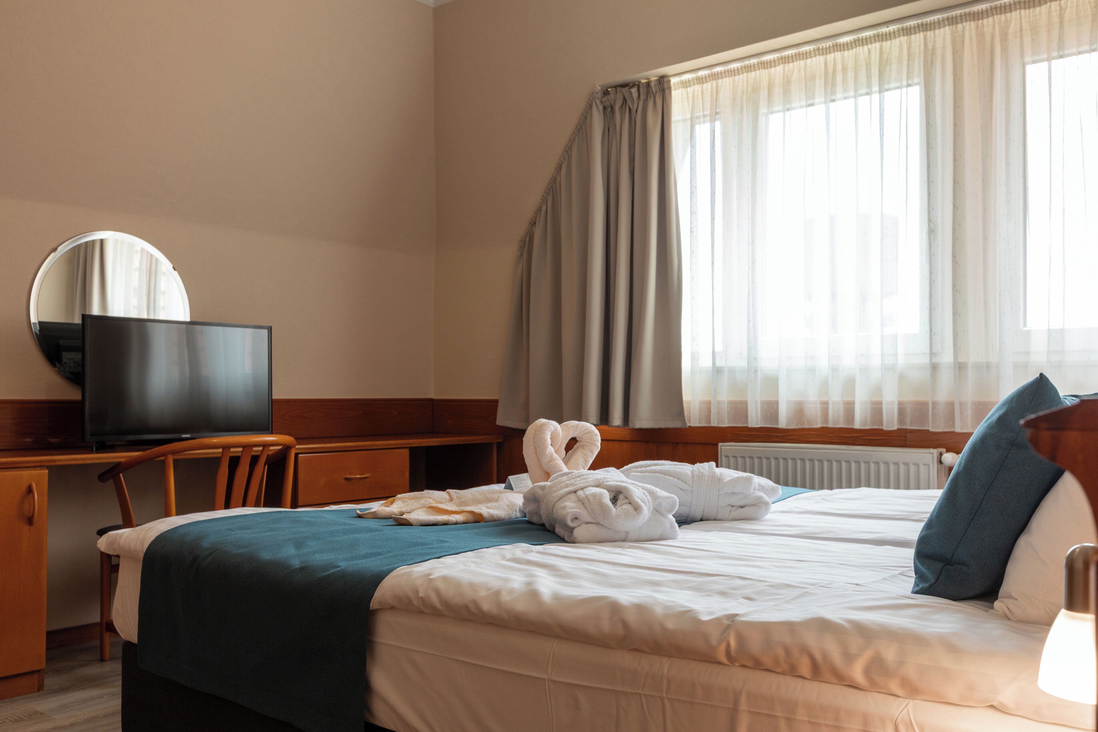 A hotel room with a bed, a desk with a television, a mirror, and a window with curtains.