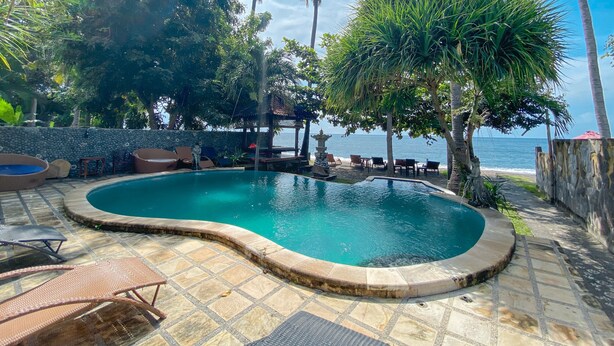 A swimming pool with lounge chairs and a stone wall, surrounded by palm trees and a view of the ocean. - Bali Bhuana Beach Cottage (Karangasem)