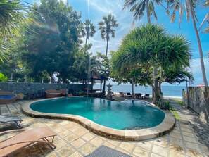 A swimming pool with lounge chairs and a stone wall, surrounded by palm trees and a view of the ocean. - Bali Bhuana Beach Cottage (Karangasem)