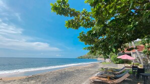 A beachside setting with lounge chairs and a table under a tree, overlooking the sea. - Bali Bhuana Beach Cottage (Karangasem)