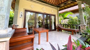 A covered outdoor seating area with wooden furniture, a tiled floor, and a view of lush greenery. - Bali Bhuana Beach Cottage (Karangasem)