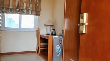 Quarto com armário de madeira, pequeno frigorífico, mesa com cadeiras e janela com cortinas.