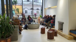 Un groupe de personnes est assis dans un hall d’entrée moderne, avec une grande fenêtre, des meubles en osier et une table en verre au centre.