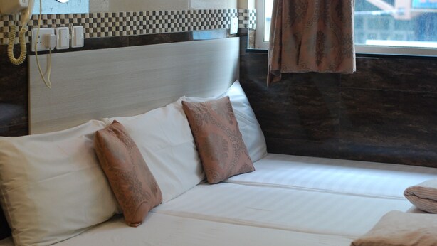 A bed with white linens and two decorative pillows, a checkered wall pattern above, and a window with curtains. - New Garden Hostel (Kowloon)