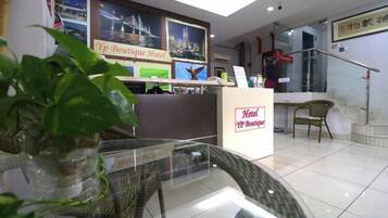 Un espace d’accueil d’hôtel comprenant un comptoir de réception, une plante en pot, des chaises en osier et un panneau indiquant le nom de l’hôtel.
