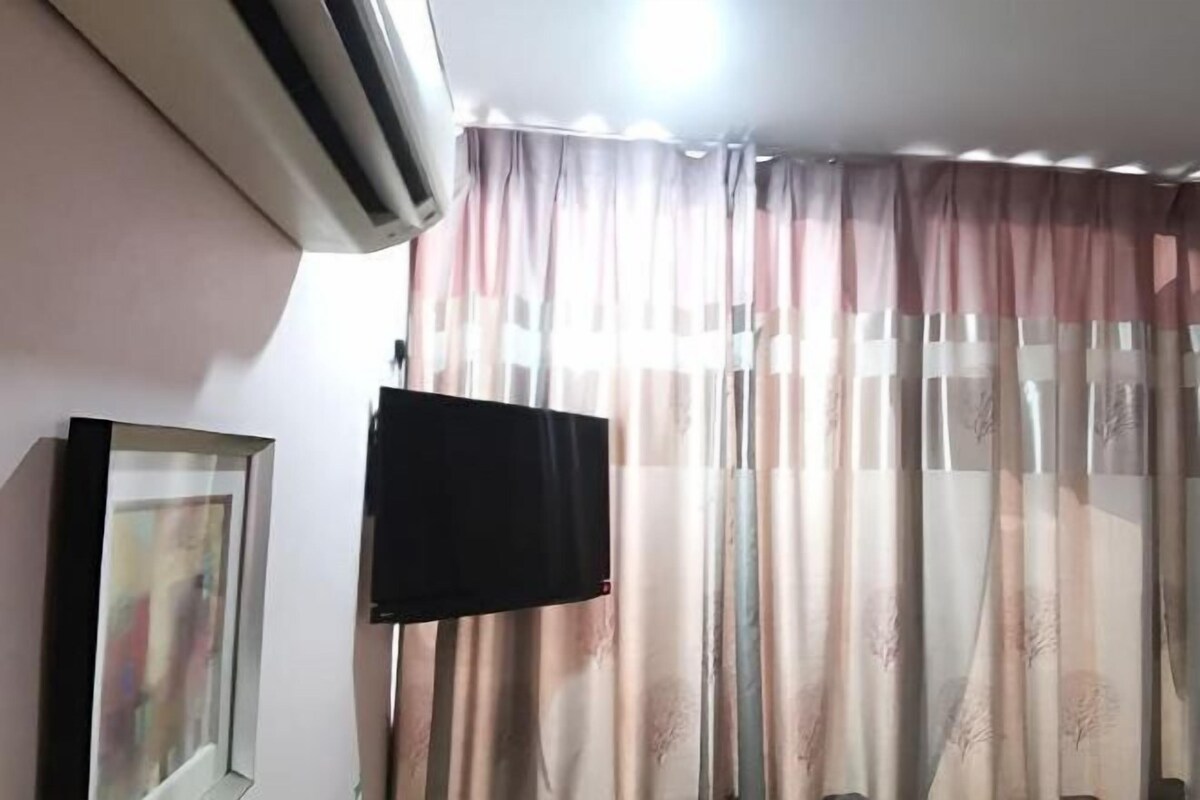 A room with a mounted television, a framed picture on the wall, and a window with sheer curtains.