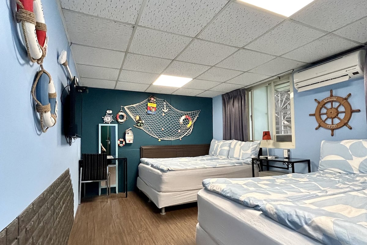 A room with two beds, a wall-mounted clock, a ship's wheel, and a window with curtains.