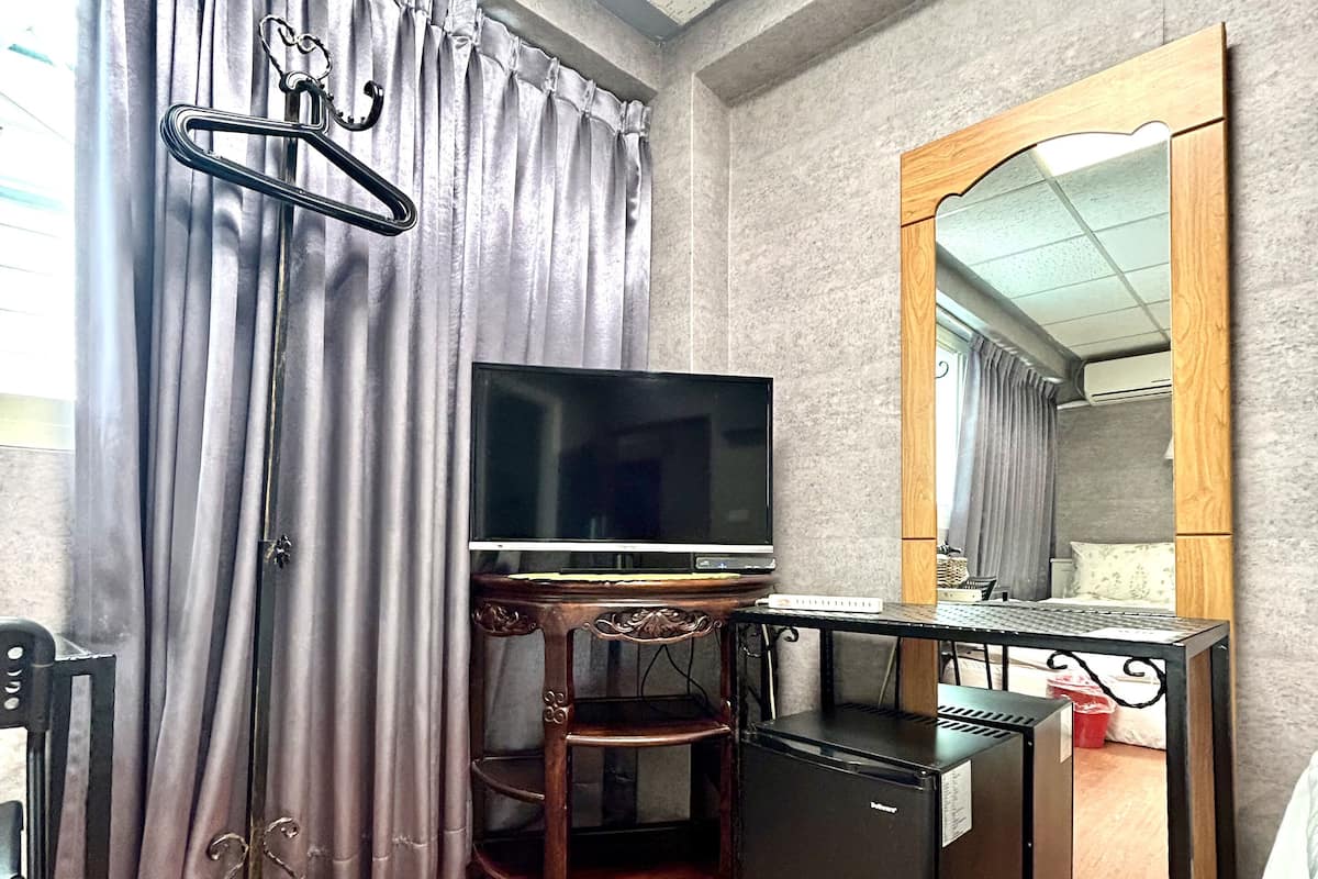 A compact hotel room with a flat-screen TV, a wooden desk, a small refrigerator, a mirror, and a bed.