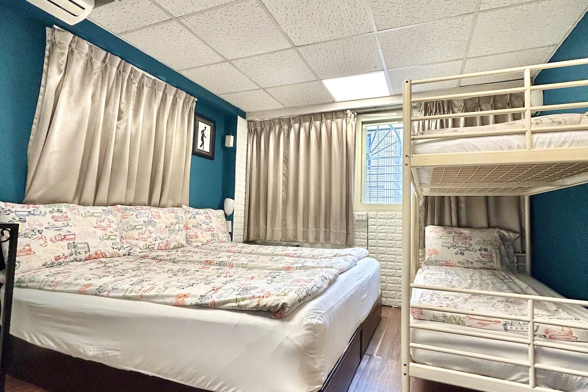 A dormitory room with a bunk bed, a single bed, wooden flooring, a window with curtains, and a ceiling with a grid pattern.