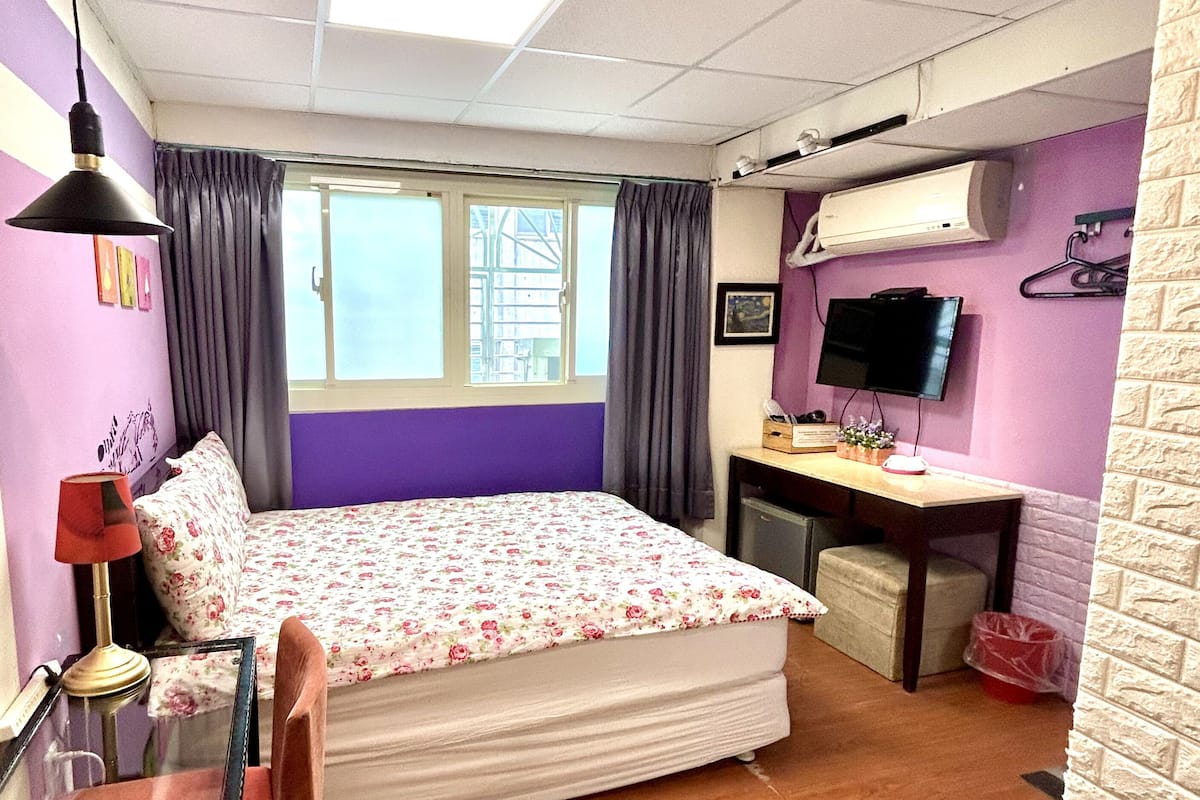 A bedroom with a bed, a desk, a chair, a TV, and a window.