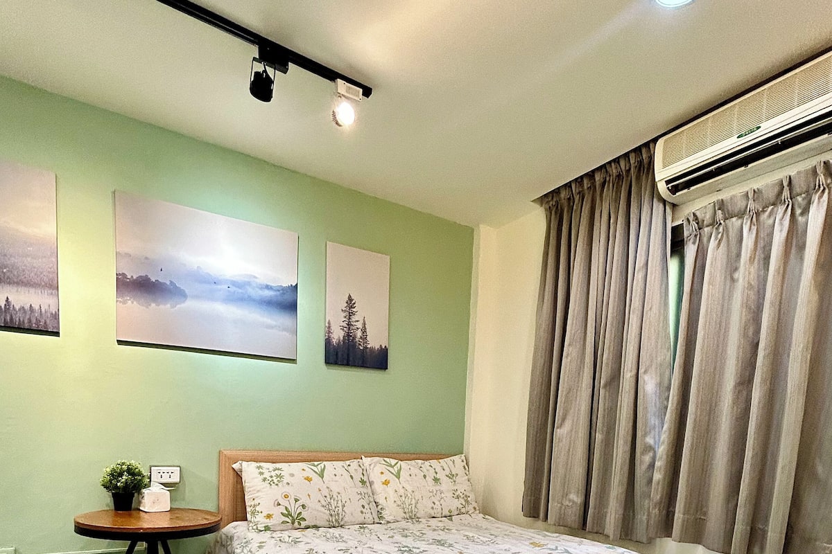A bedroom with a large bed, a small table with a plant, and artwork on the wall.