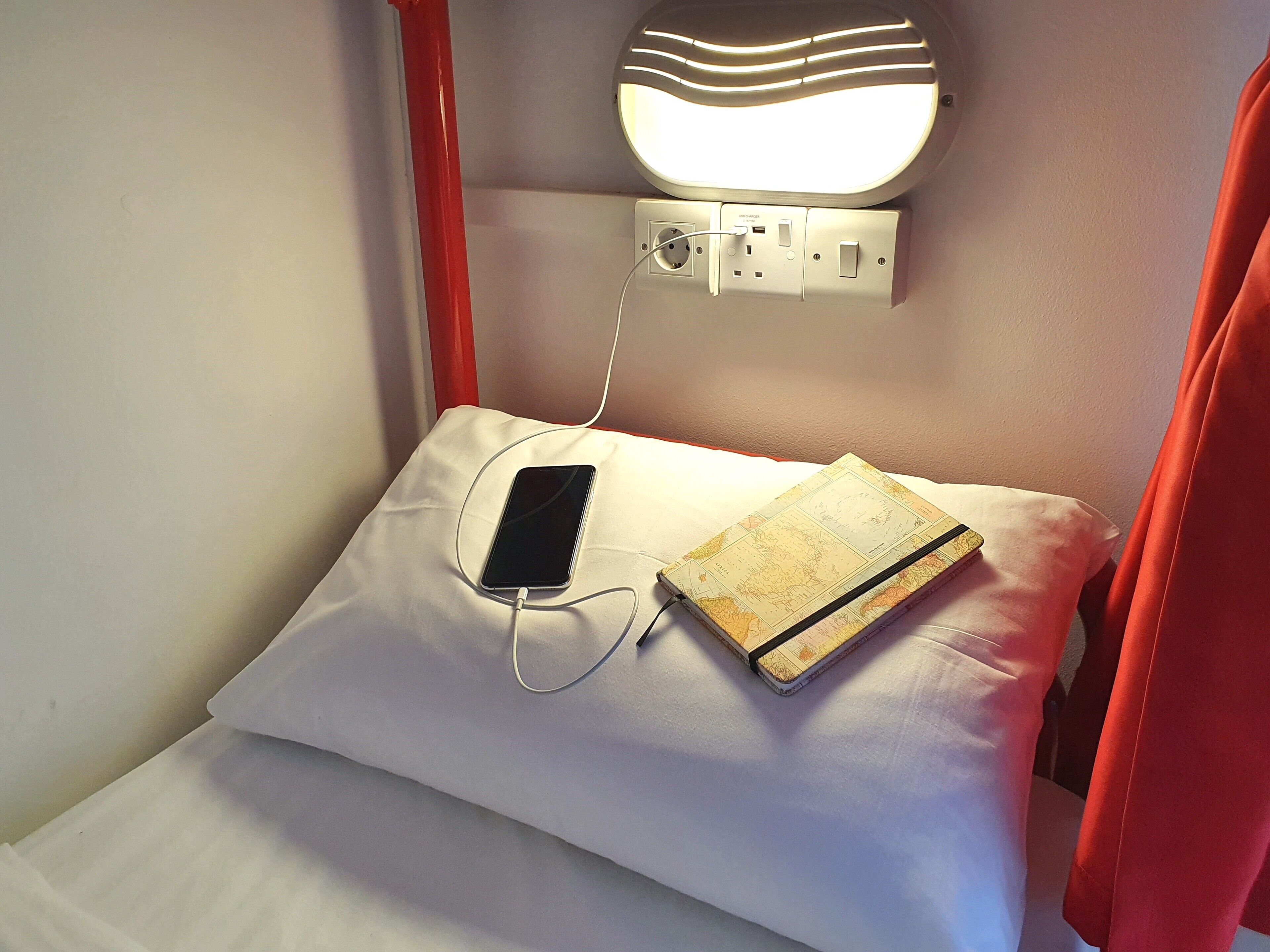 A bed with a white pillow and a map, a smartphone connected to a charger, and a wall-mounted light fixture.