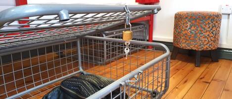 A room with a metal cart containing a black bag, a wooden floor, a red chair, and a bed with a white headboard.