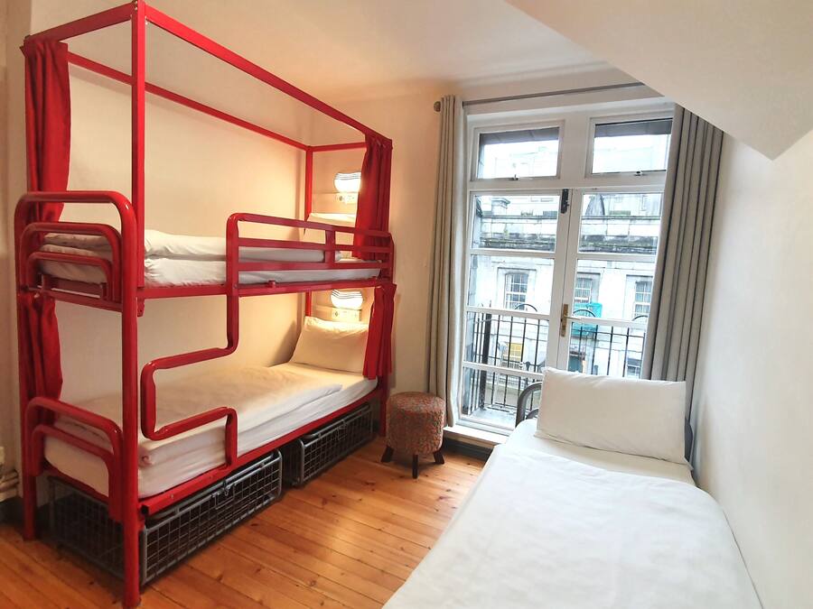 A room with a red bunk bed, a single bed, a window with a view of buildings, and a small stool.