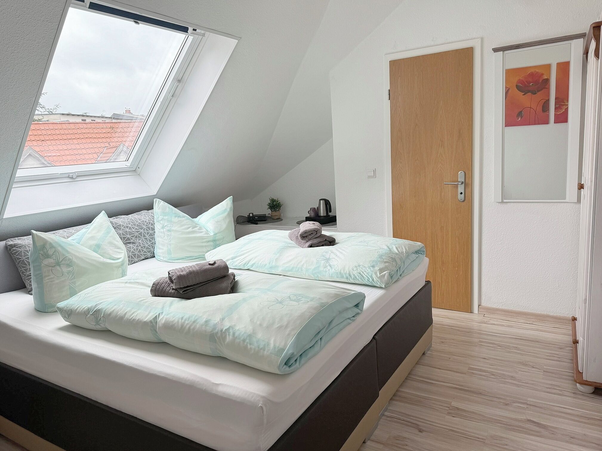Un dormitorio con techo inclinado, una cama con varias almohadas, una puerta de madera y una pequeña mesa con una planta.