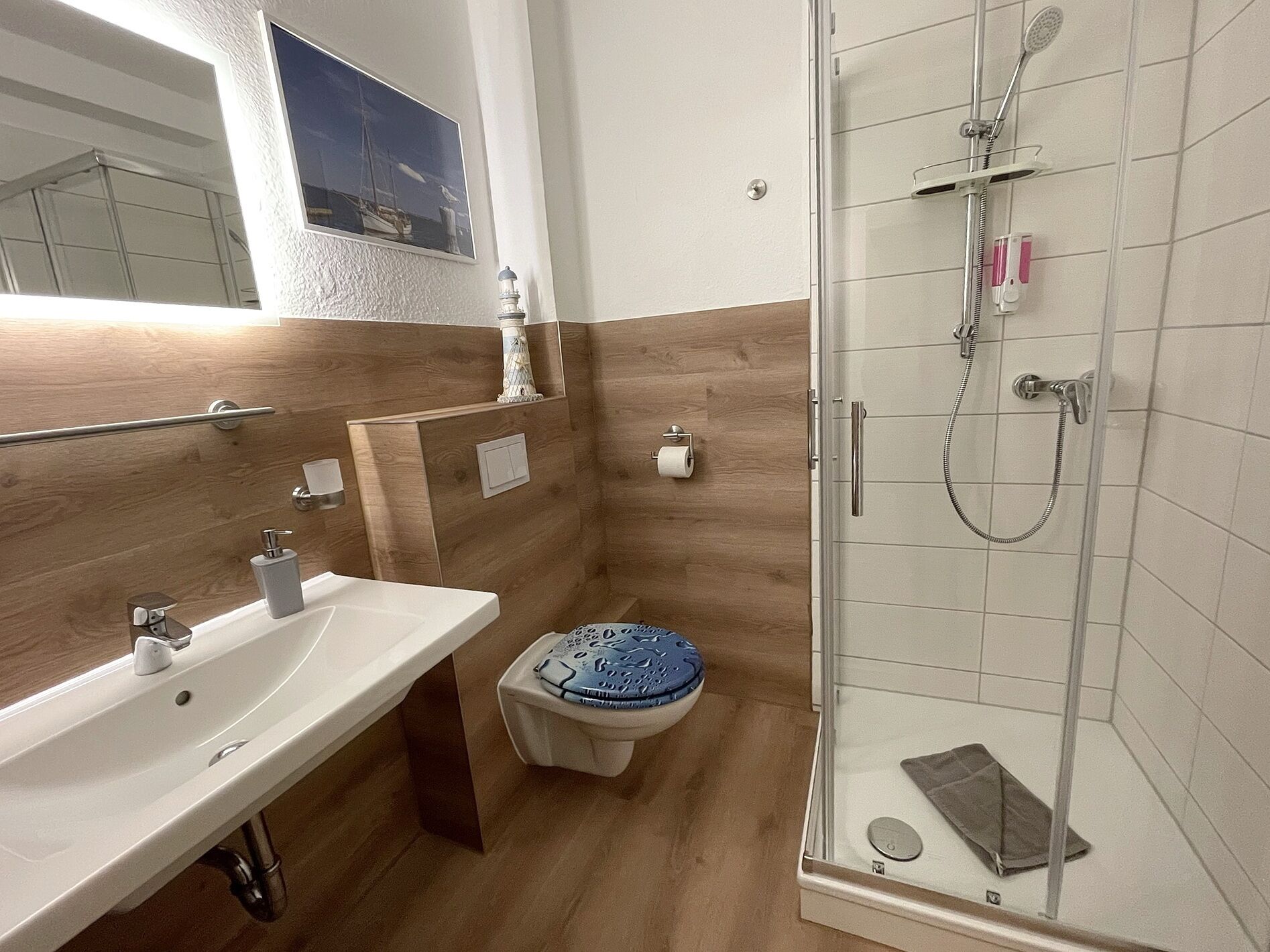 Une salle de bain avec un lavabo blanc, des toilettes avec un abattant bleu, une douche avec une porte en verre et un panneau mural en bois.
