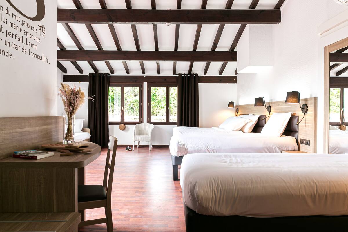 A hotel room with two beds, a desk with a chair, a vase with dried flowers, and large windows with curtains.