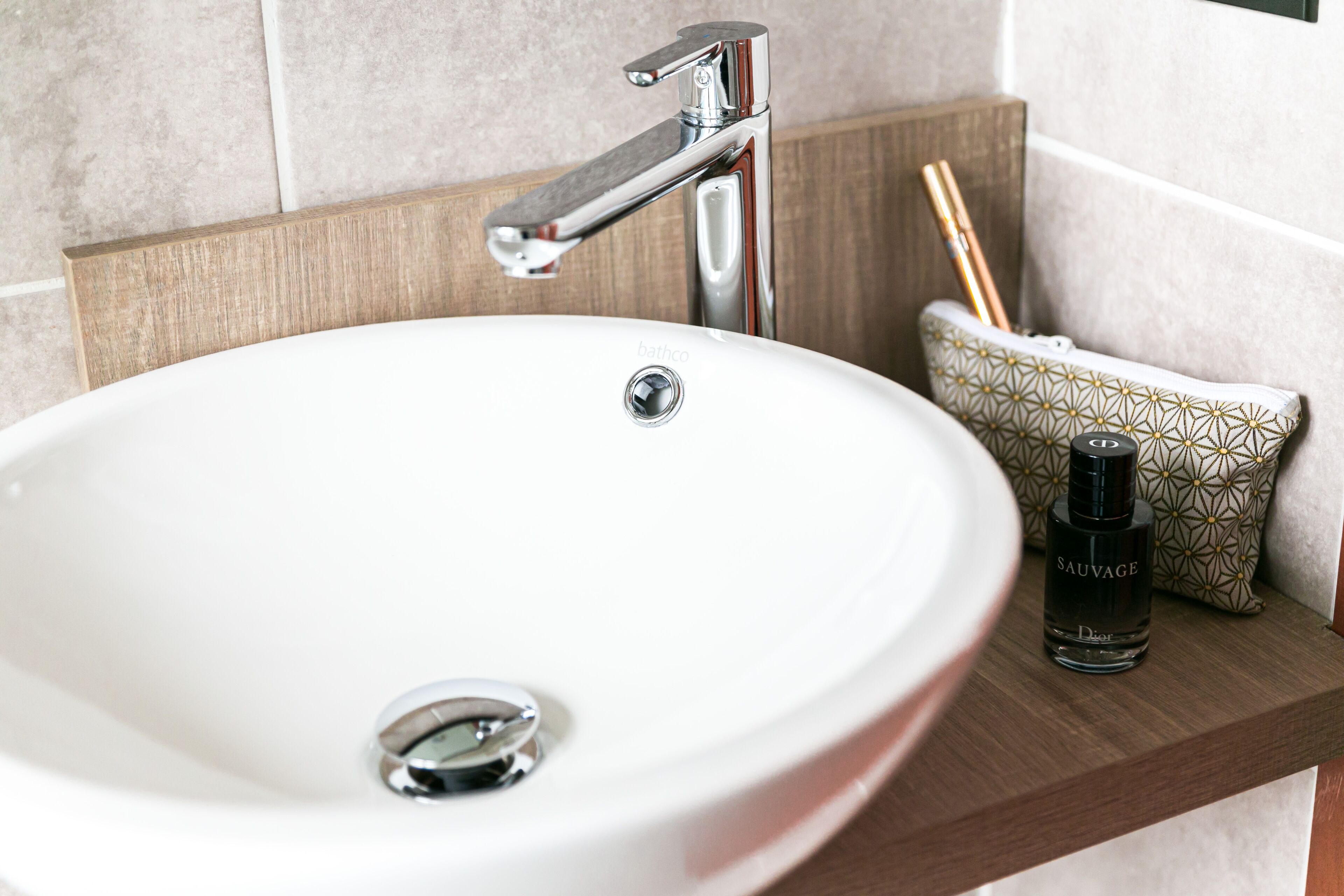 Un lavandino da bagno bianco con rubinetteria cromata, una bottiglia di profumo Sauvage e un cuscino decorato su una mensola in legno.