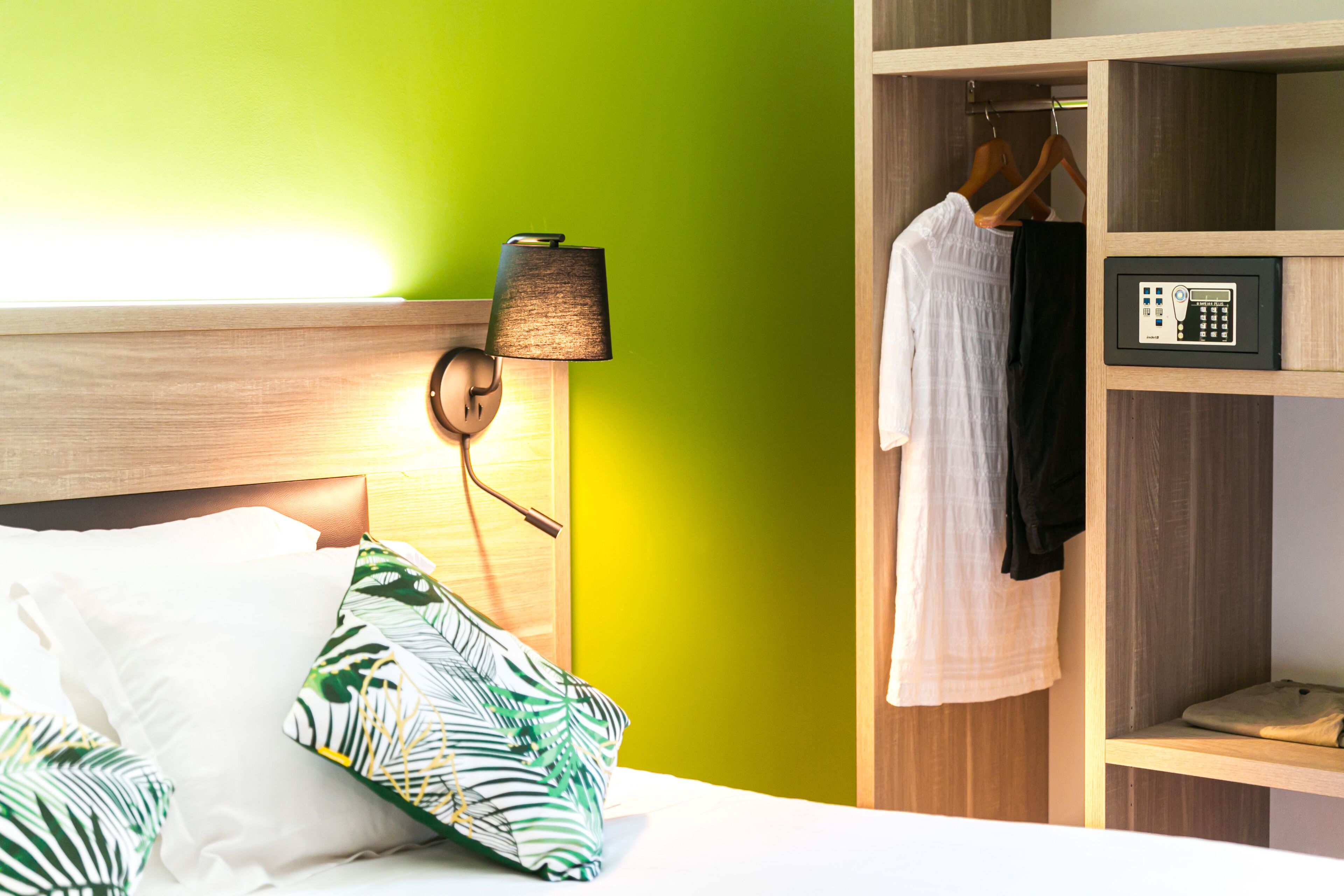 A hotel room with a bed, a nightstand with a lamp, a wardrobe with hangers, and a control panel.