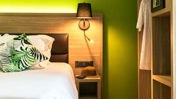 Una cama bien hecha con ropa blanca y un cojín con estampado de hojas tropicales. Una lámpara con pantalla negra está montada en la pared sobre la cama.