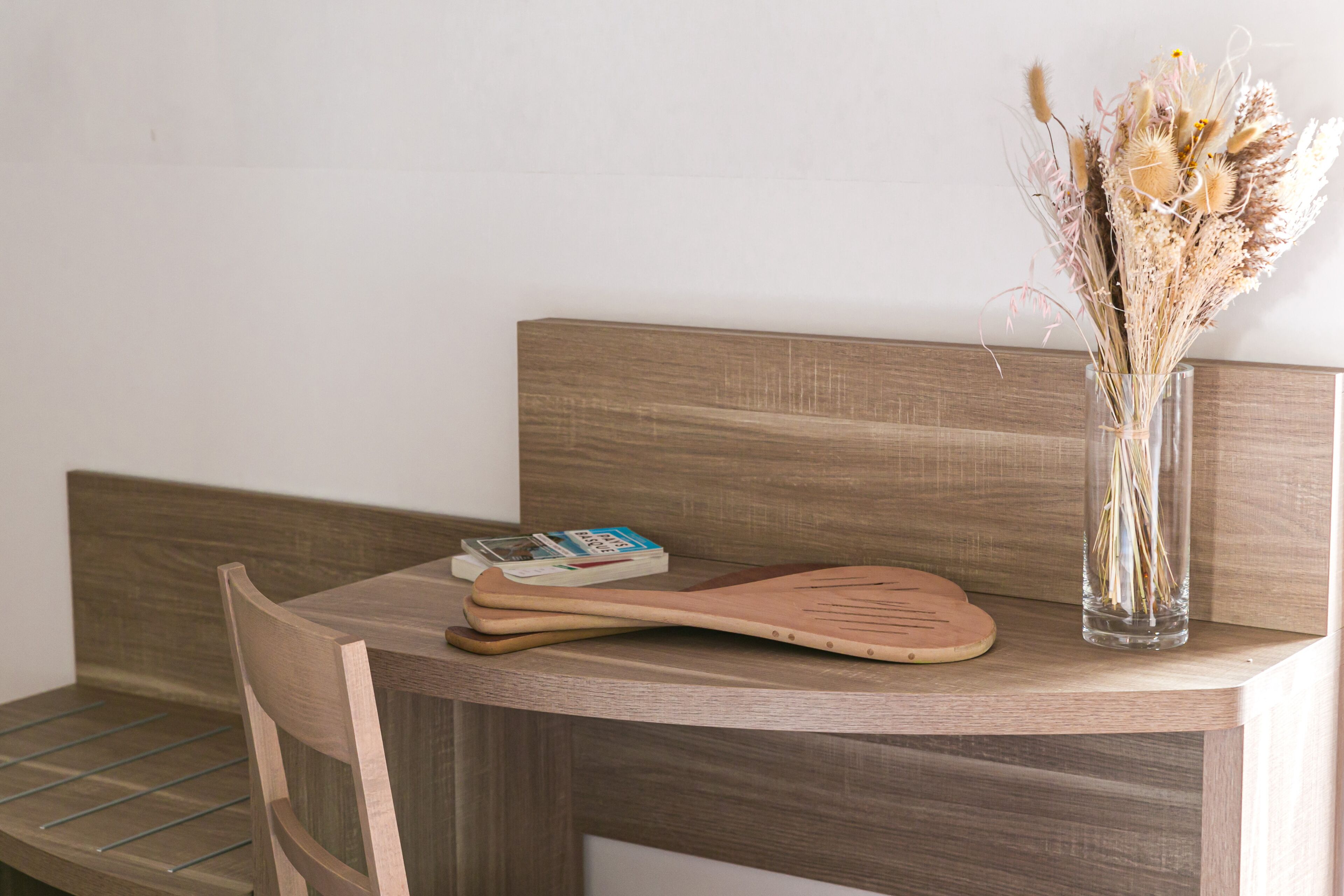 A wooden table with a wooden spatula, a book, and a vase with dried flowers.
