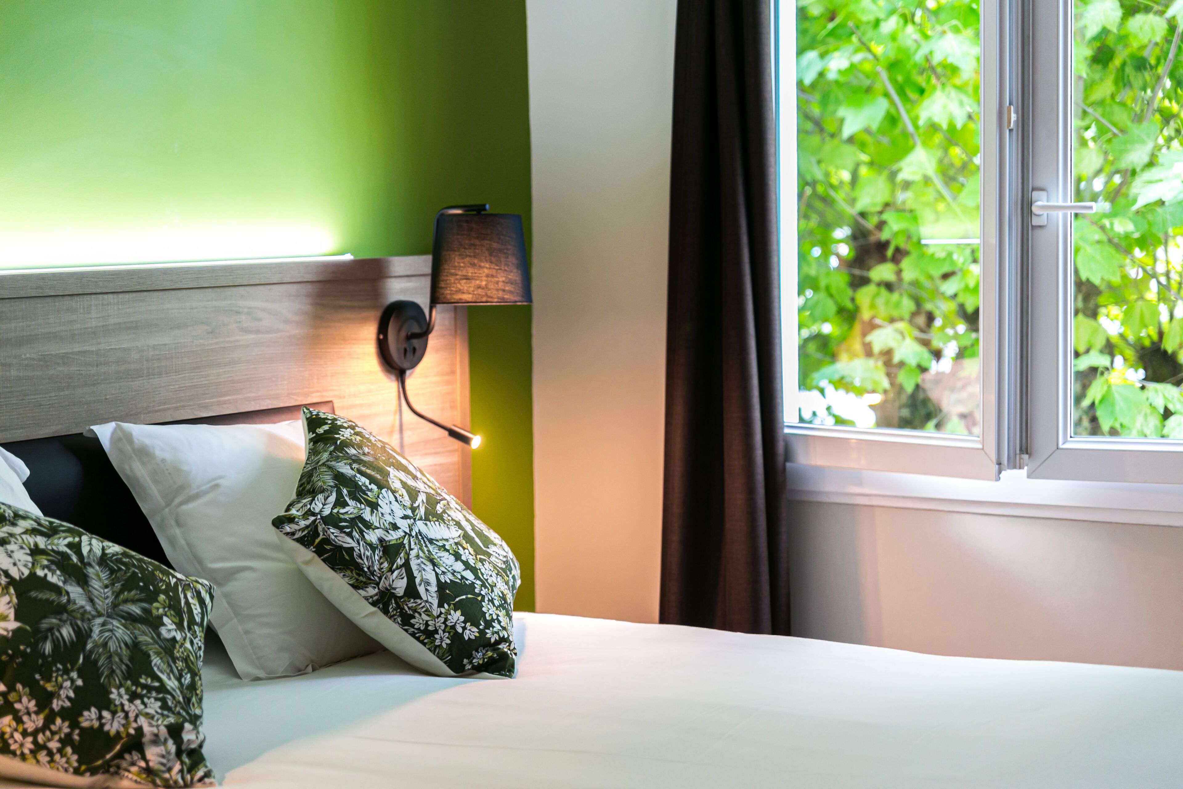 Habitación de hotel con cama, paredes verdes, ventana con vistas a vegetación y una lámpara fijada a la pared.