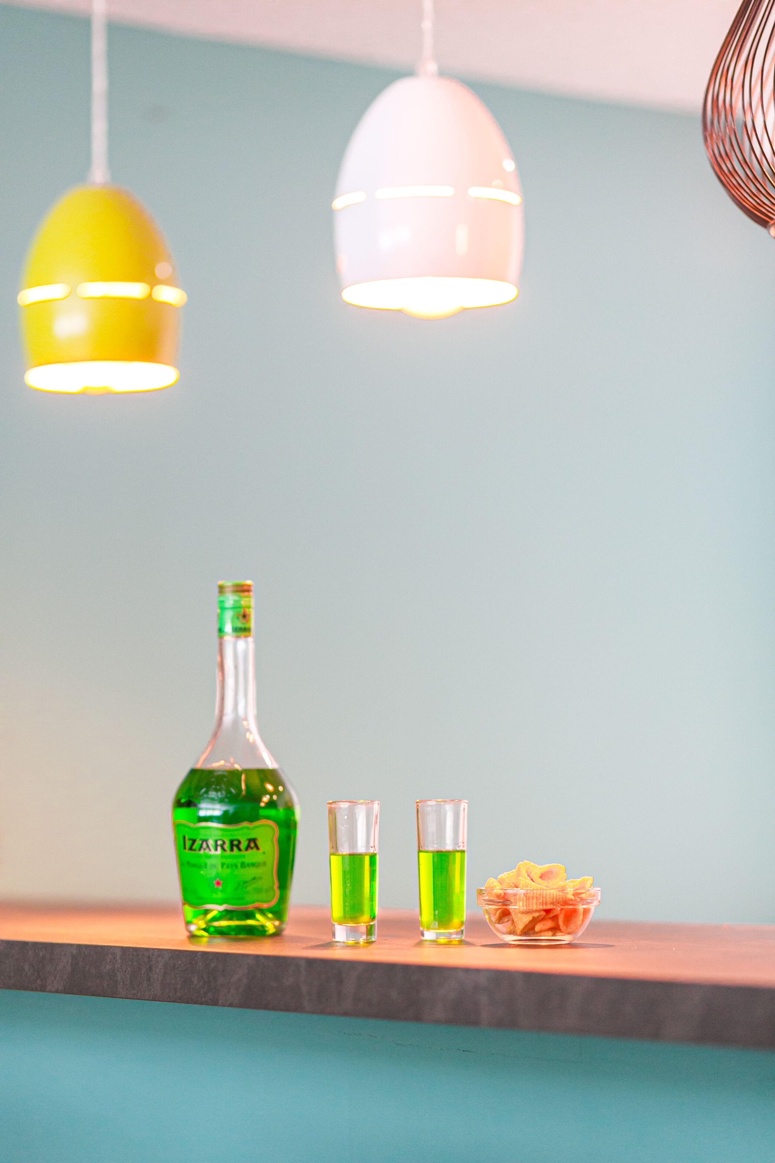 A bottle of liquor, two shot glasses, and a bowl of snacks on a wooden surface with hanging pendant lights.