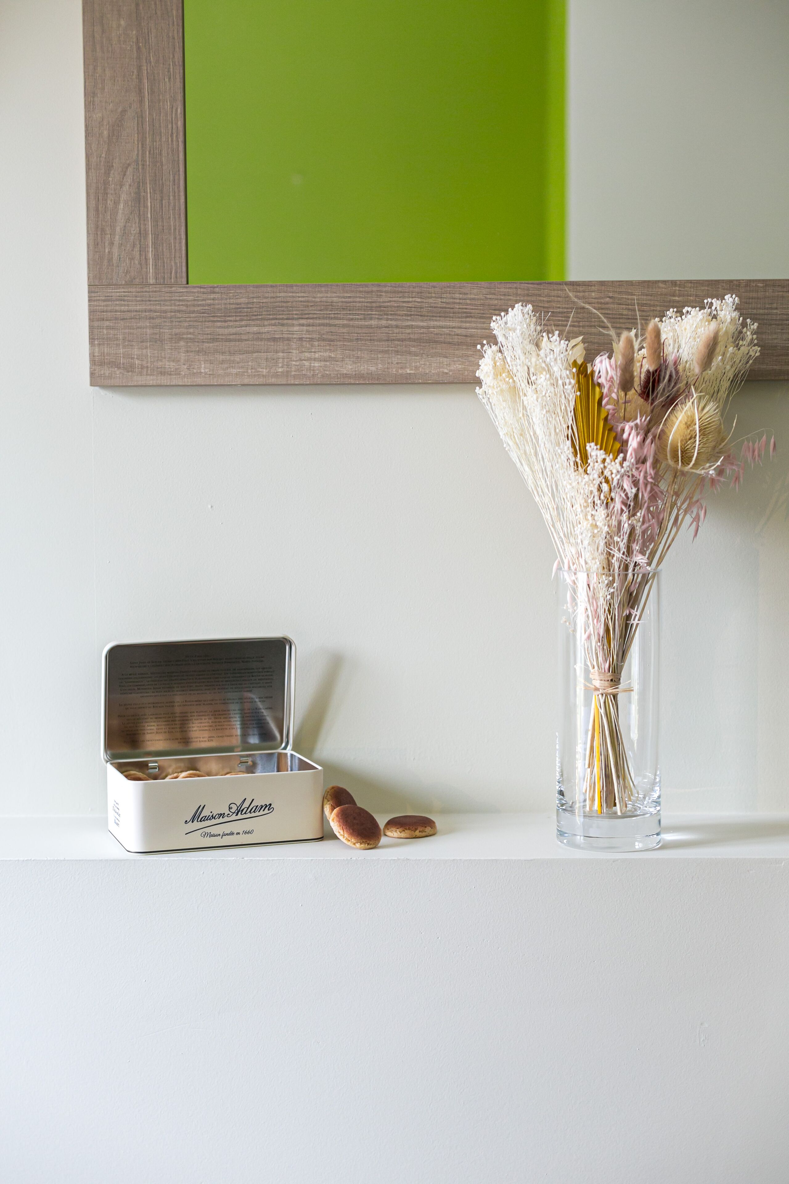 Una mesa blanca con una lata de metal con la etiqueta "Maison Adam" y un florero de vidrio con flores secas.