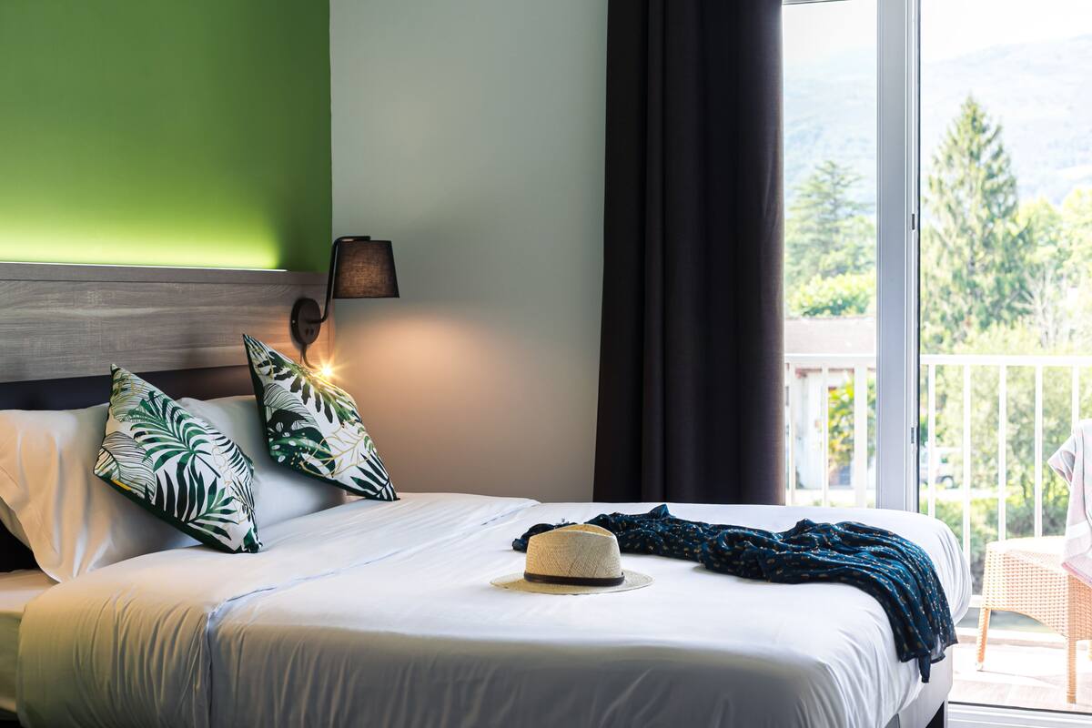 A hotel room with a bed, a hat, and a view of greenery outside.