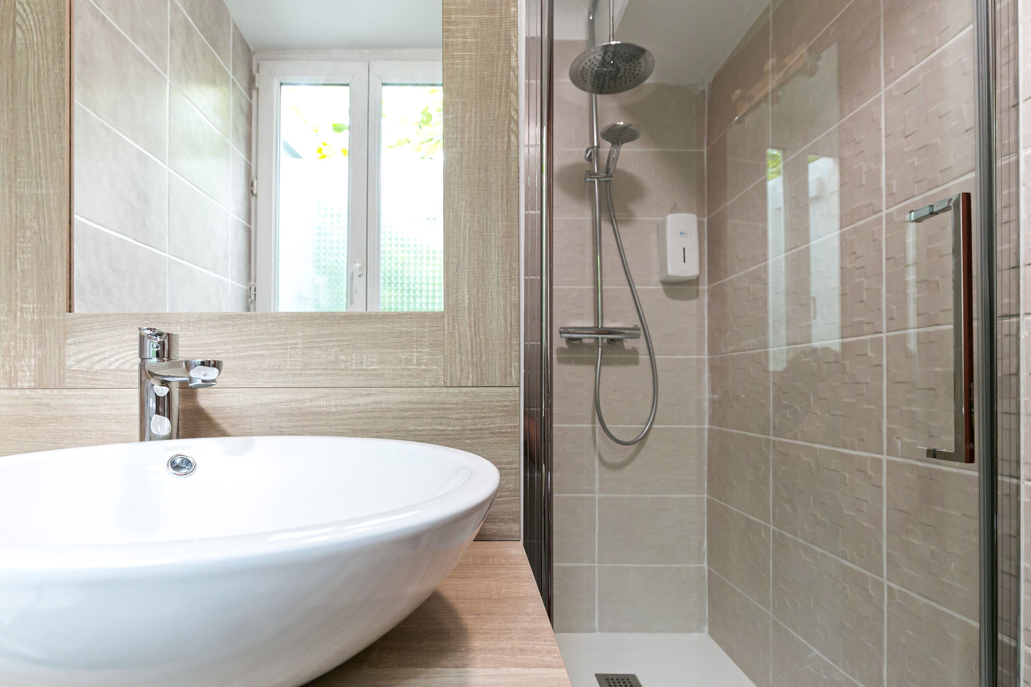 Une salle de bain moderne avec un grand lavabo blanc, une douche en verre et une fenêtre qui laisse entrer la lumière naturelle.