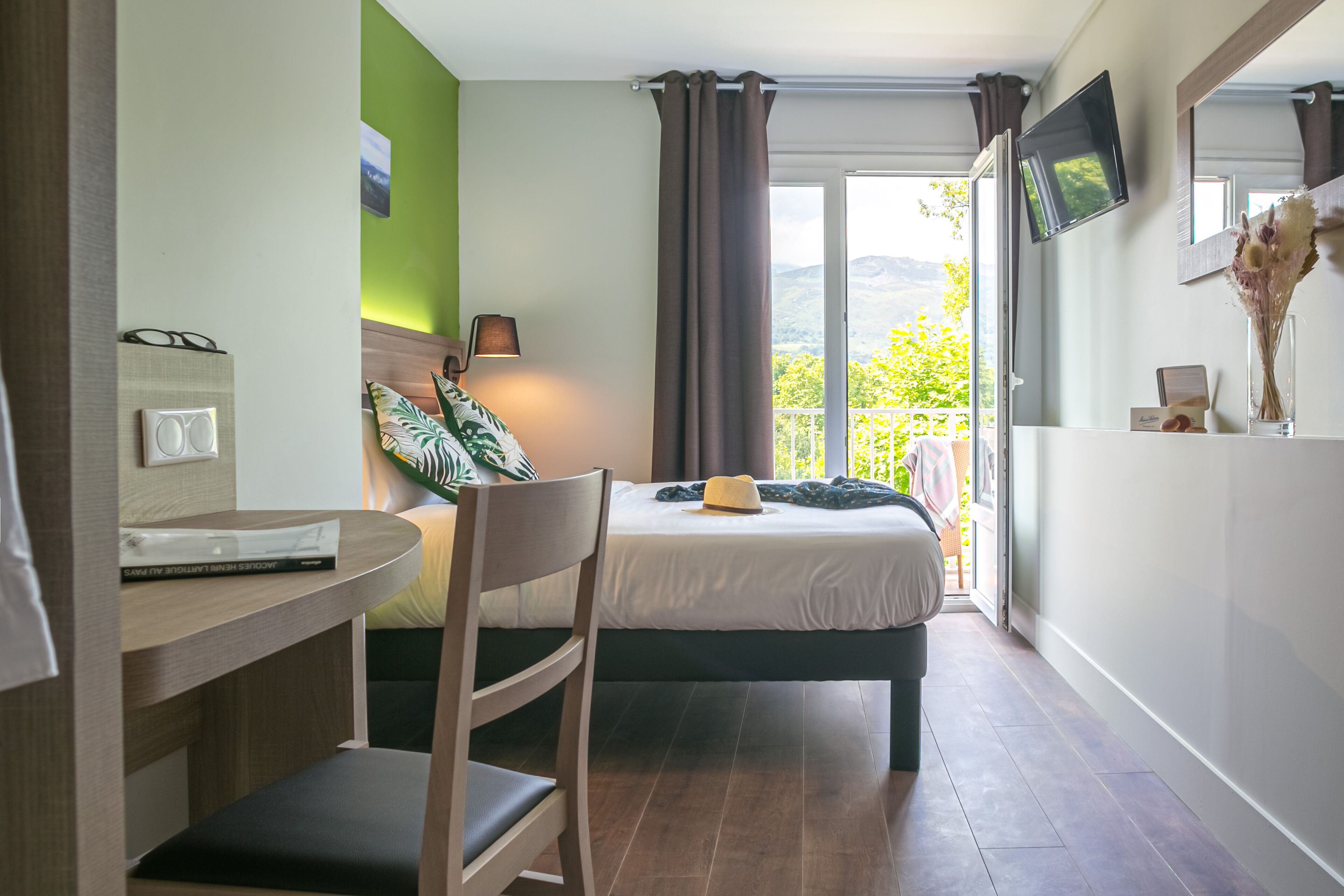 A hotel room with a bed, a desk with a chair, a TV mounted on the wall, and a view of the outdoors.