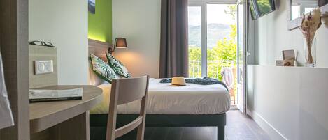 A hotel room with a bed, a desk with a chair, a TV mounted on the wall, and a view of the outdoors.