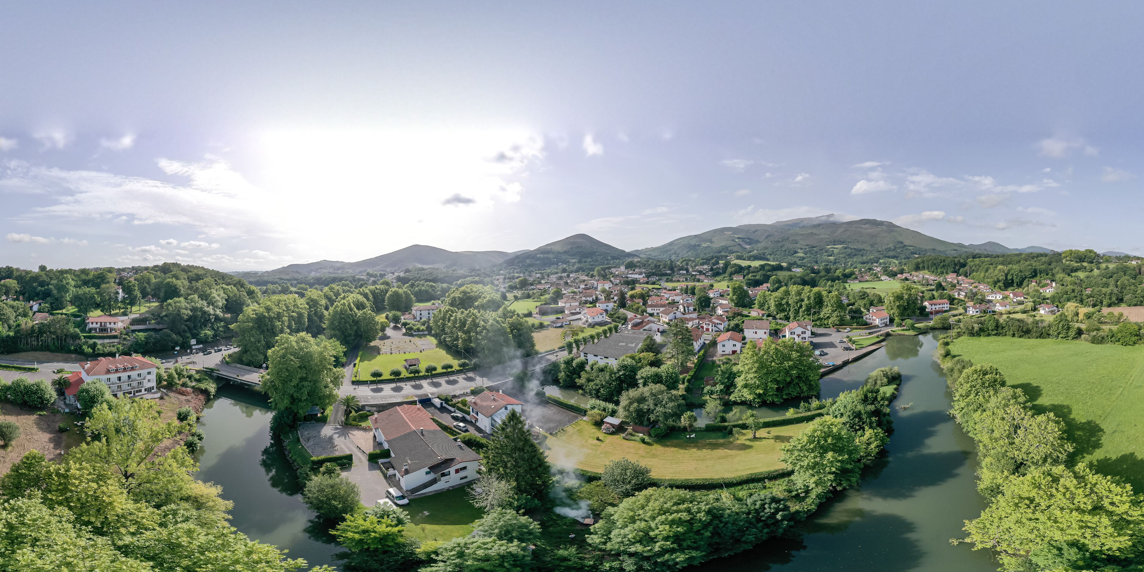 An aerial view of a riverside town with residential buildings, a bridge, and surrounding greenery.
